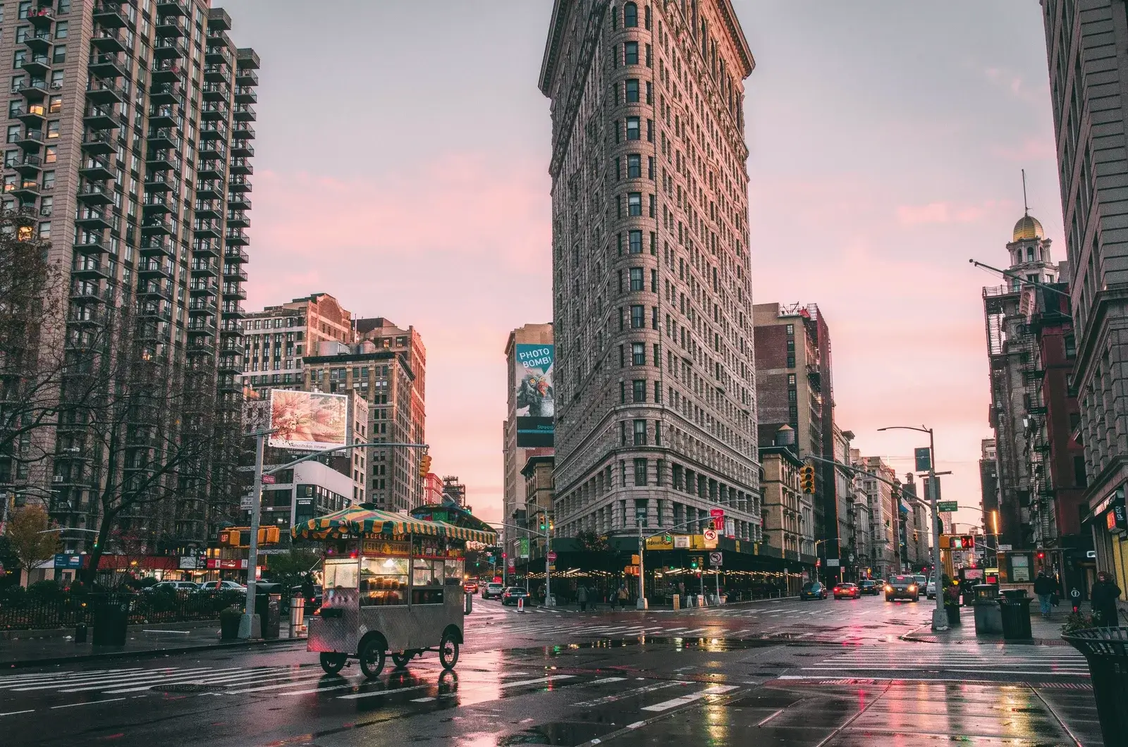 The narrow wedge of the Flatiron Building at dawn under a pink sky, a coffee cart with a striped awning on the wet corner in front of it