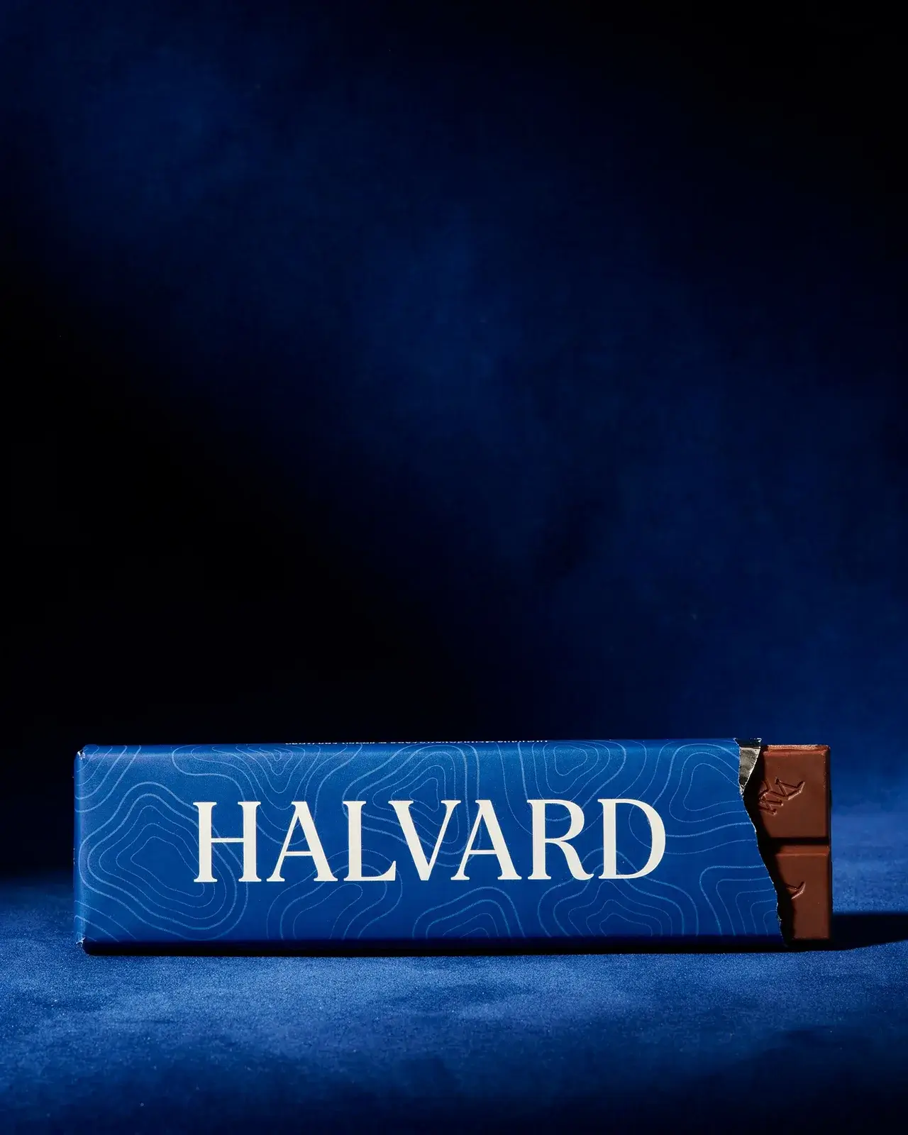 A Halvard bar in a cobalt contour-line wrapper on deep blue velvet along the bottom of the frame, two squares of chocolate showing, the dark velvet above left empty for a headline