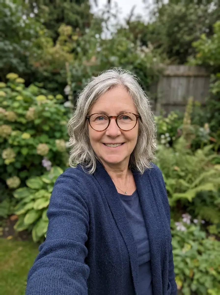 A phone selfie of a woman around sixty with wavy grey hair, round tortoiseshell glasses and a navy cardigan in a garden