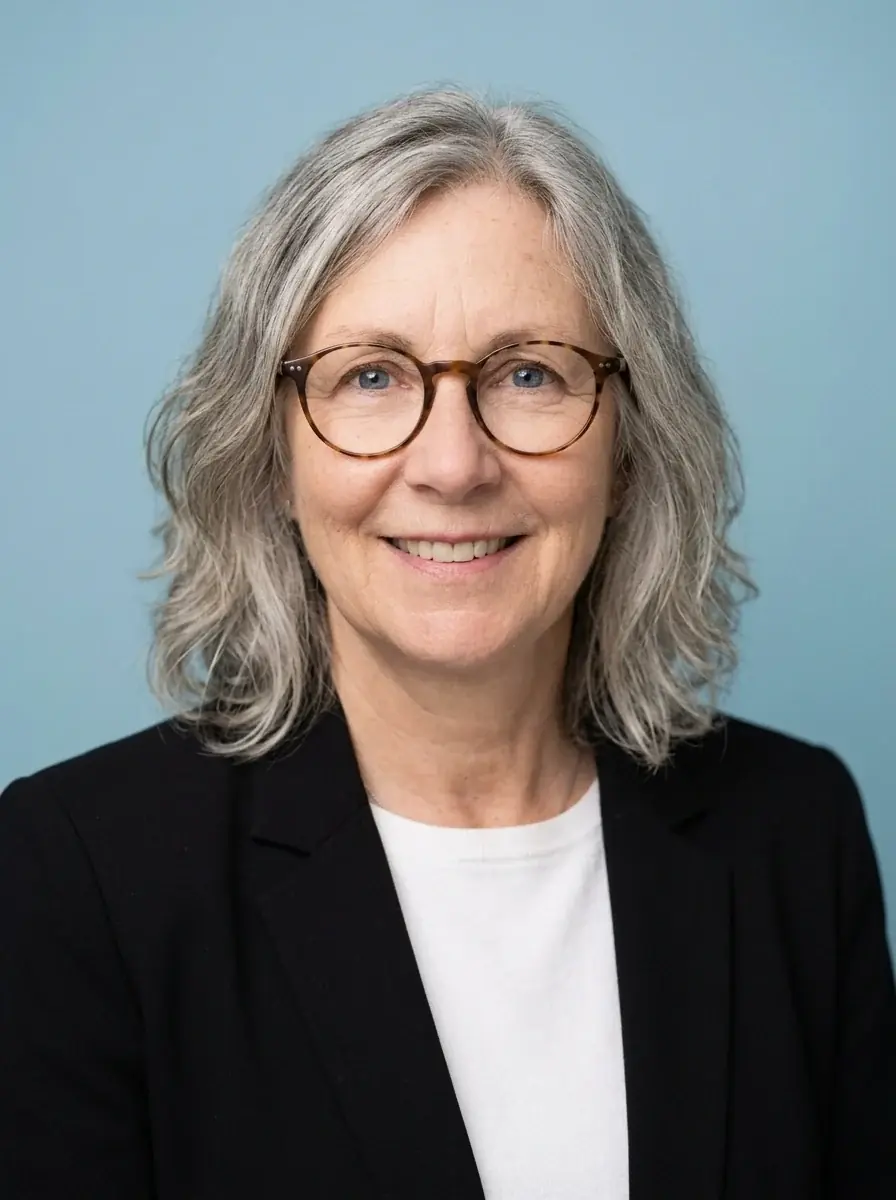 A studio headshot of a woman with wavy grey hair and tortoiseshell glasses in a black blazer against a soft blue backdrop
