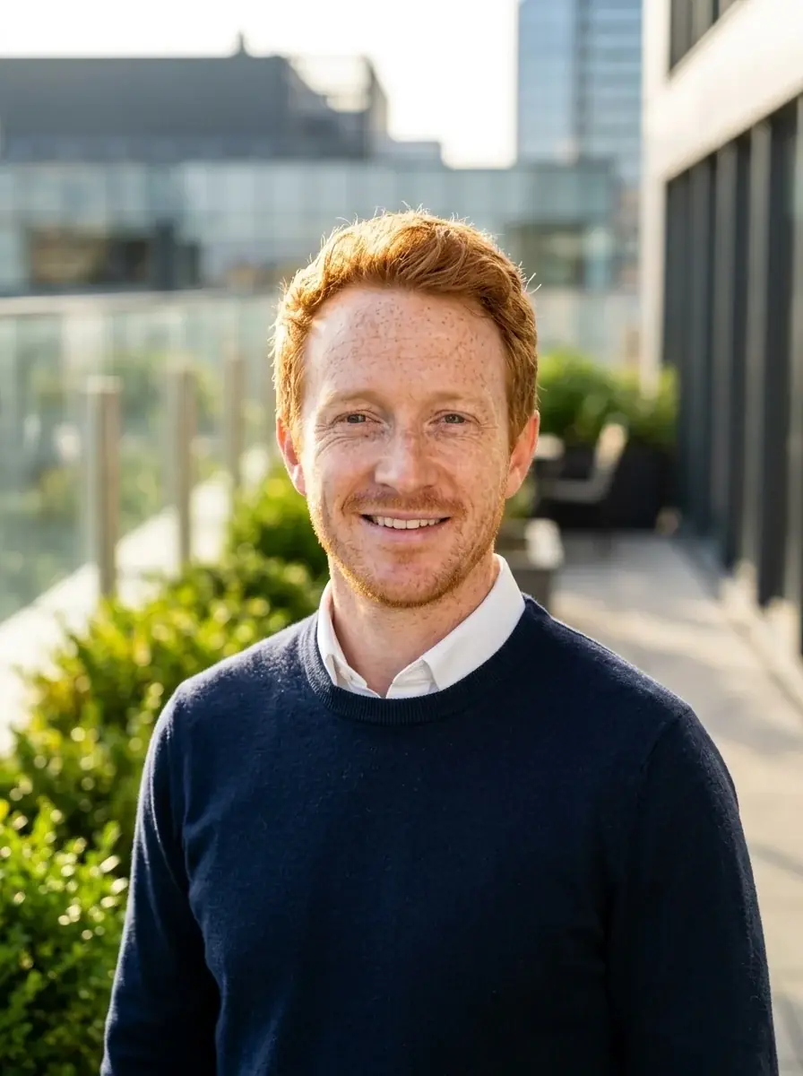 An outdoor headshot of a freckled man with ginger hair in a navy sweater over a white collar on a sunny office terrace