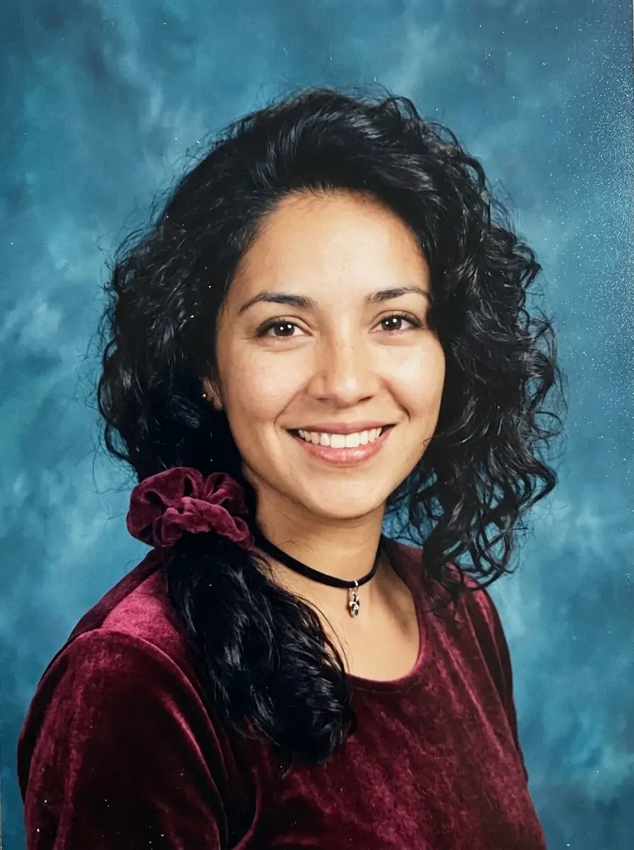 A 90s yearbook portrait of a woman with big curly hair and a scrunchie in a burgundy velvet top and choker against a teal backdrop