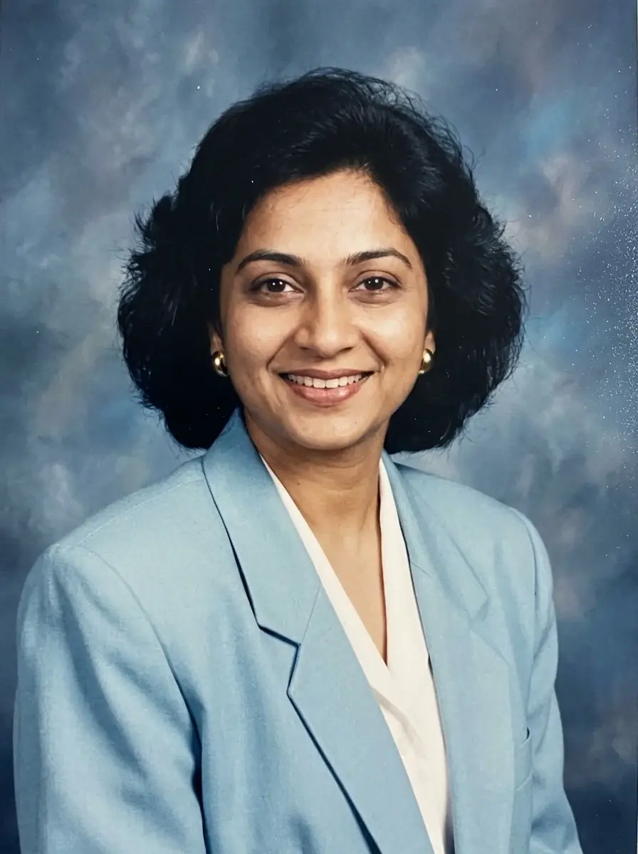 A 90s yearbook portrait of a woman with a feathered bob in a pastel blue blazer with shoulder pads against a mottled blue backdrop