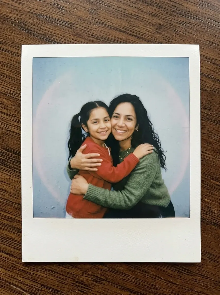 A Polaroid of a woman with long wavy hair in a green sweater hugging herself as a girl with pigtails in a red cardigan