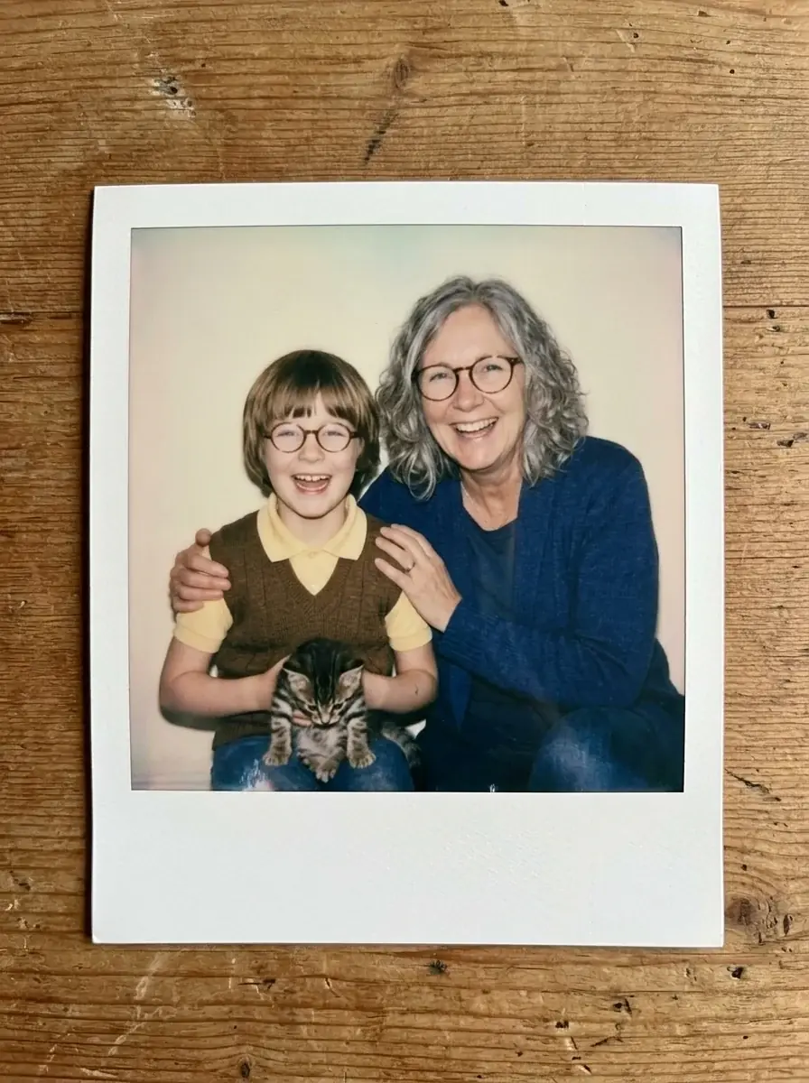 A Polaroid of a grey-haired woman in glasses and a navy cardigan laughing beside herself as a girl in glasses holding a kitten
