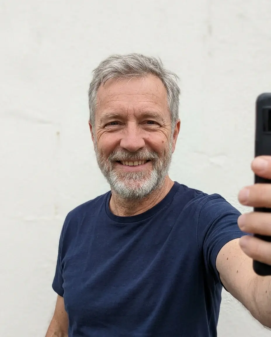 A phone selfie of a grinning older man with short grey hair and a grey beard in a navy t-shirt, against a white wall