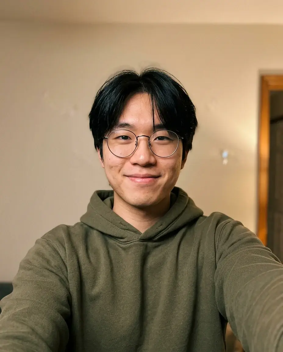 A phone selfie of a young man with black hair and round wire glasses in an olive hoodie, in a room lit by a warm lamp