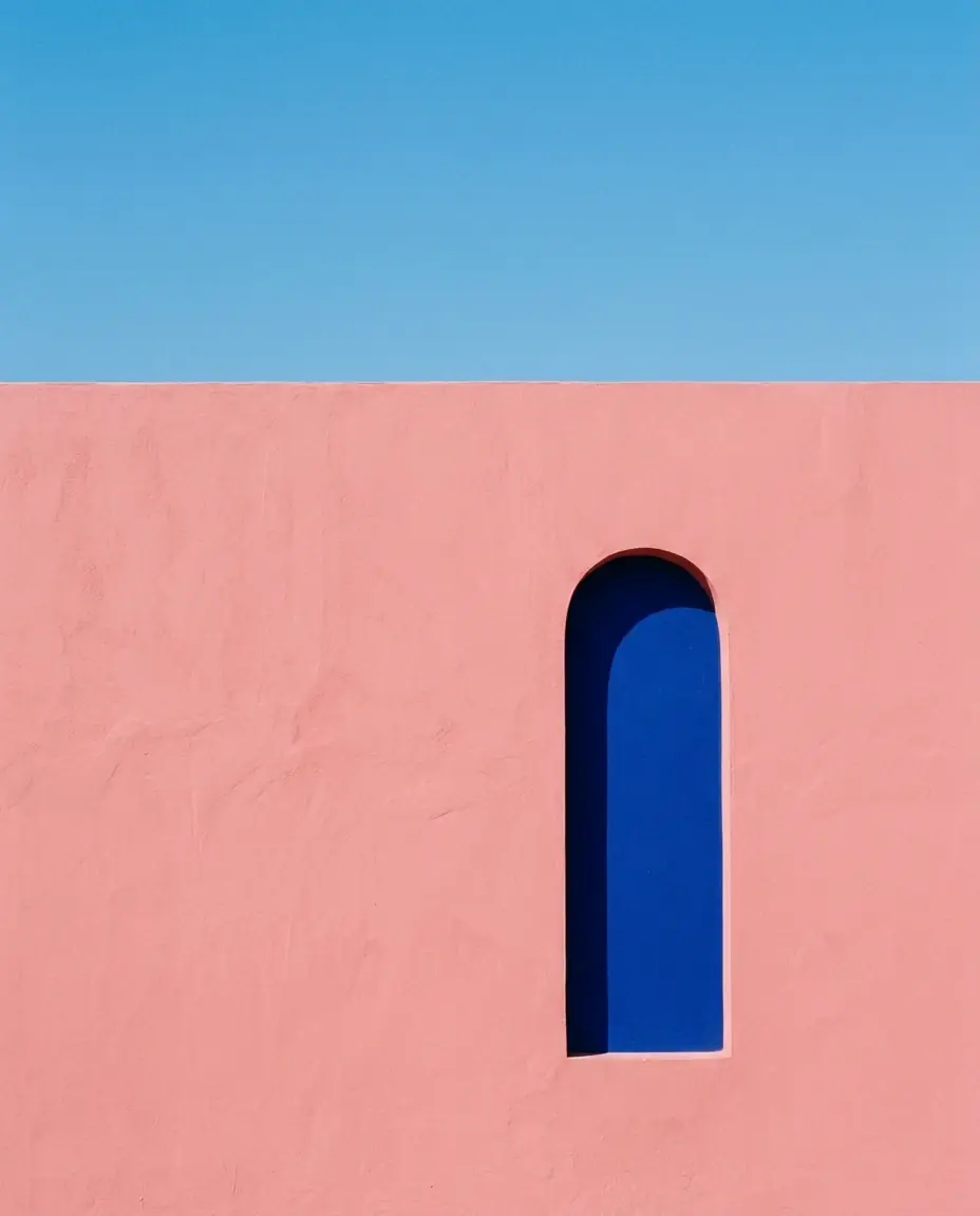 A pink stucco wall with one tall arched opening painted deep ultramarine, under a clear blue sky, the top of the wall a straight line across the upper third