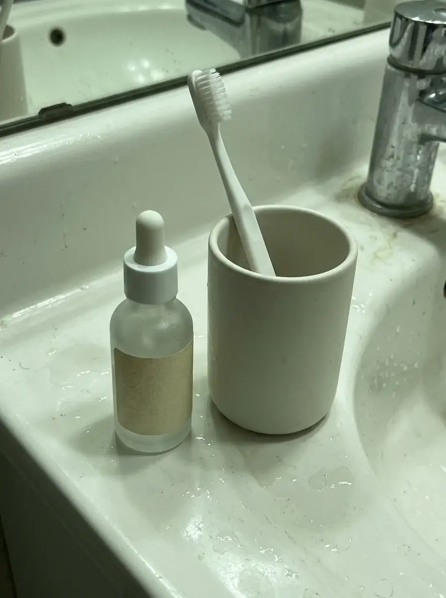 A generated phone photo of a frosted serum dropper bottle with a blank paper label on a white bathroom sink beside a toothbrush cup, dim light with a green cast and water spots on the basin
