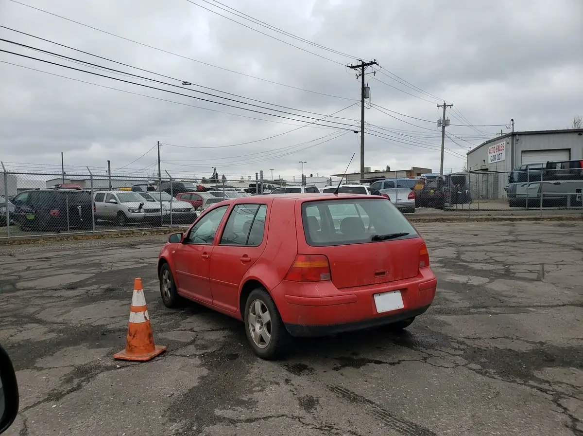 The same red hatchback after Ideogram Edit: the cars on both sides gone, empty asphalt around it, the hatchback, cone and fence unchanged