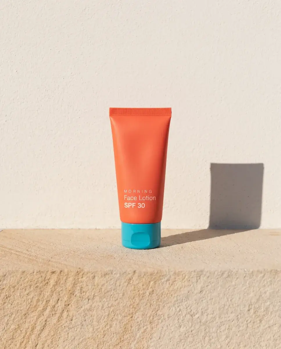 The orange face lotion tube on the same sandstone ledge and chalk-white wall, its shadow to the right in the same hard morning sun