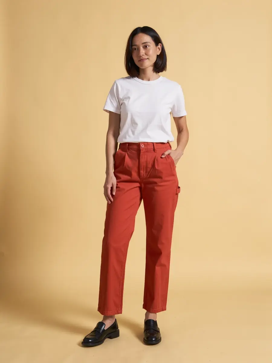 A woman wearing the tomato red trousers with a white T-shirt and black loafers, one hand in a pocket, on a warm yellow paper backdrop