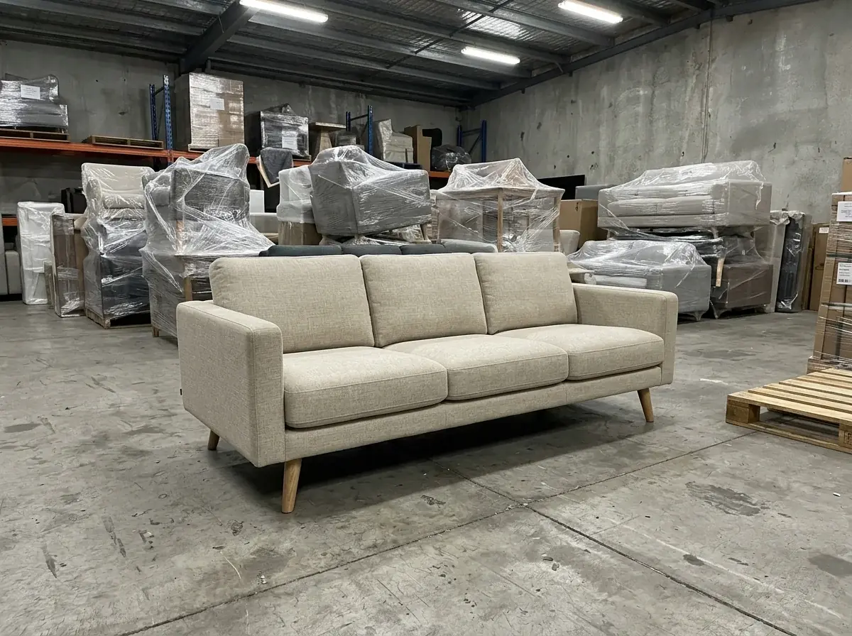 A generated phone photo of an oat linen three-seat sofa with tapered oak legs on a concrete warehouse floor, plastic-wrapped furniture and a pallet behind it under fluorescent tubes