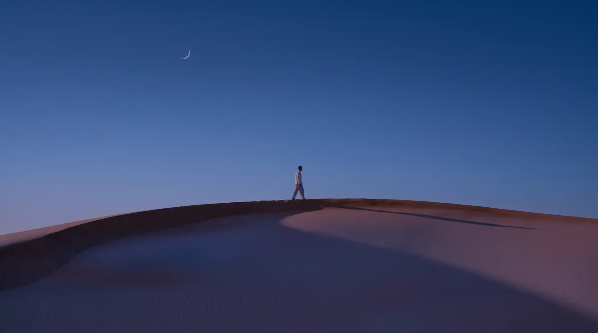 The same dune at blue hour for dark mode: a deep indigo sky with a thin crescent moon on the left, the walker and the ridge unchanged