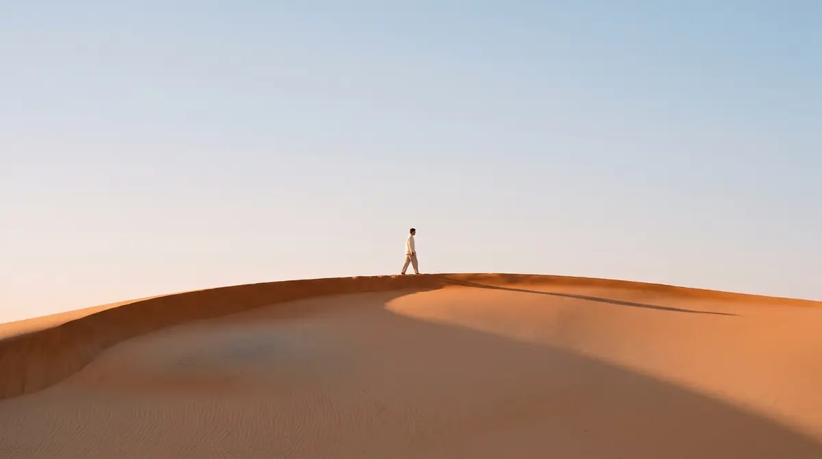 A small walker in a cream shirt on the crest of an apricot dune in early morning, the ridge curving across the lower third, open pale sky above