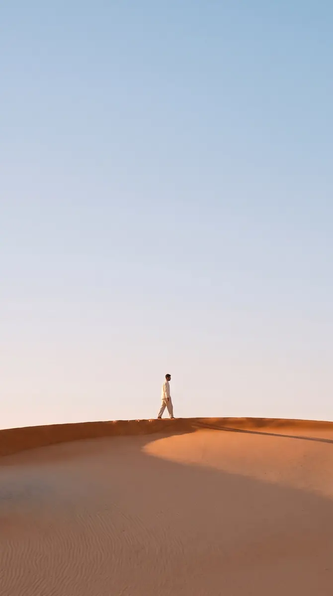 The dune recomposed as a tall phone wallpaper: the walker small on the crest in the lower third, open pale blue sky above