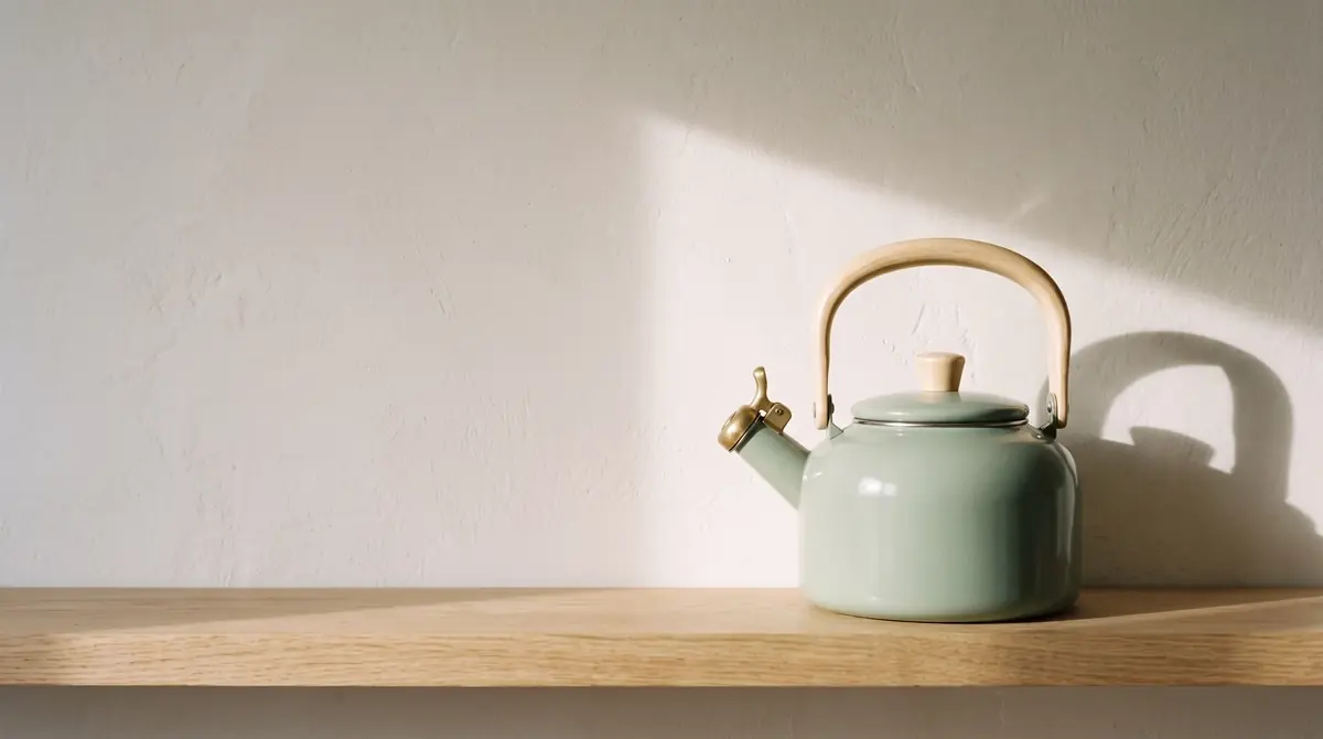 The same kettle alone on a pale oak shelf against warm white plaster in the right third, morning sun casting its shadow on the wall, the left half calm and empty