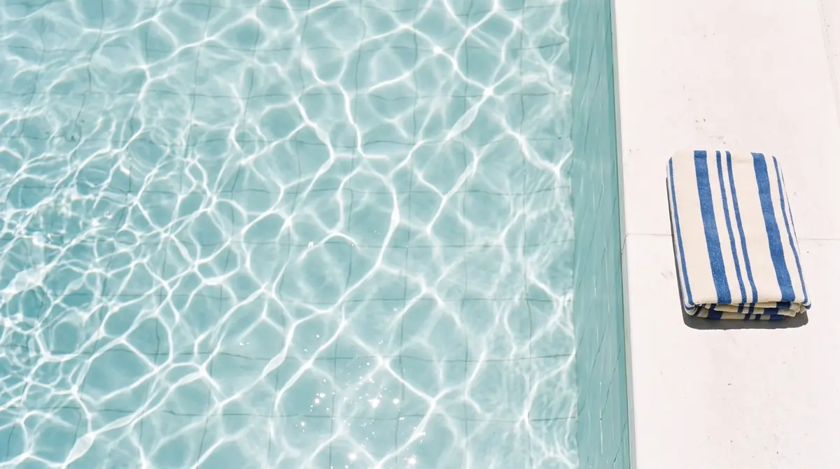 Looking down into a pool at noon: a net of white caustic lines on pale aqua tiles, a limestone edge on the right with one folded cream and cobalt striped towel