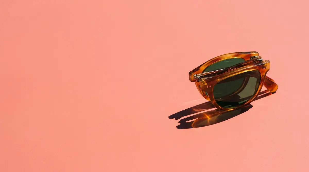 Folded amber tortoiseshell sunglasses with bottle green lenses in the right third of a coral pink plane, a crisp shadow with a warm amber glow in it, the left half empty
