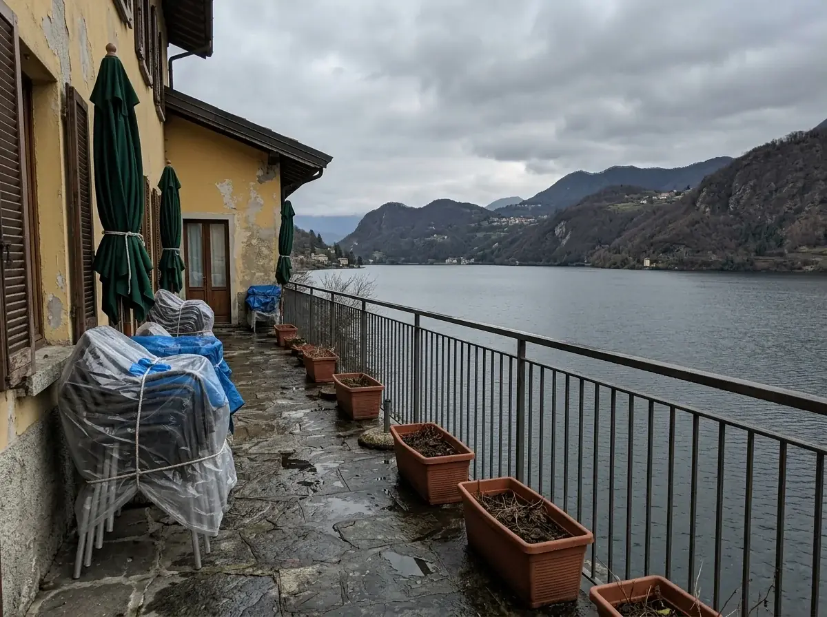 A generated phone photo of a hotel terrace above a lake in early spring: chairs stacked under plastic, folded green parasols, empty planters, wet stone floor and a gray sky