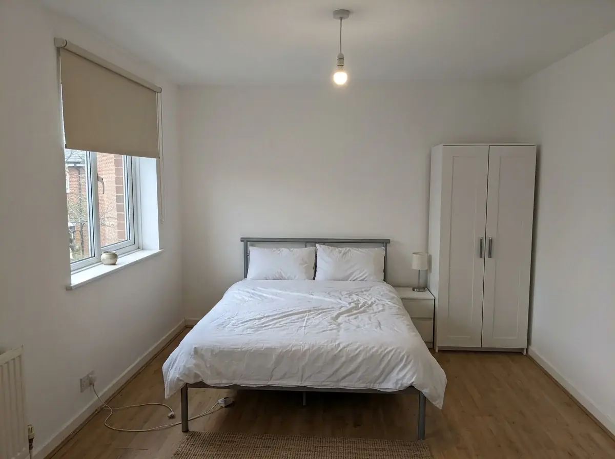 A generated phone photo of a plain rental bedroom: white walls, a metal double bed with a white duvet, a bare bulb, a white wardrobe and a window with a roller blind
