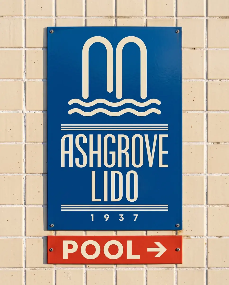 A blue enamel sign on a cream tiled wall with the arch mark and the ASHGROVE LIDO 1937 wordmark in cream, a red panel below reading POOL with an arrow
