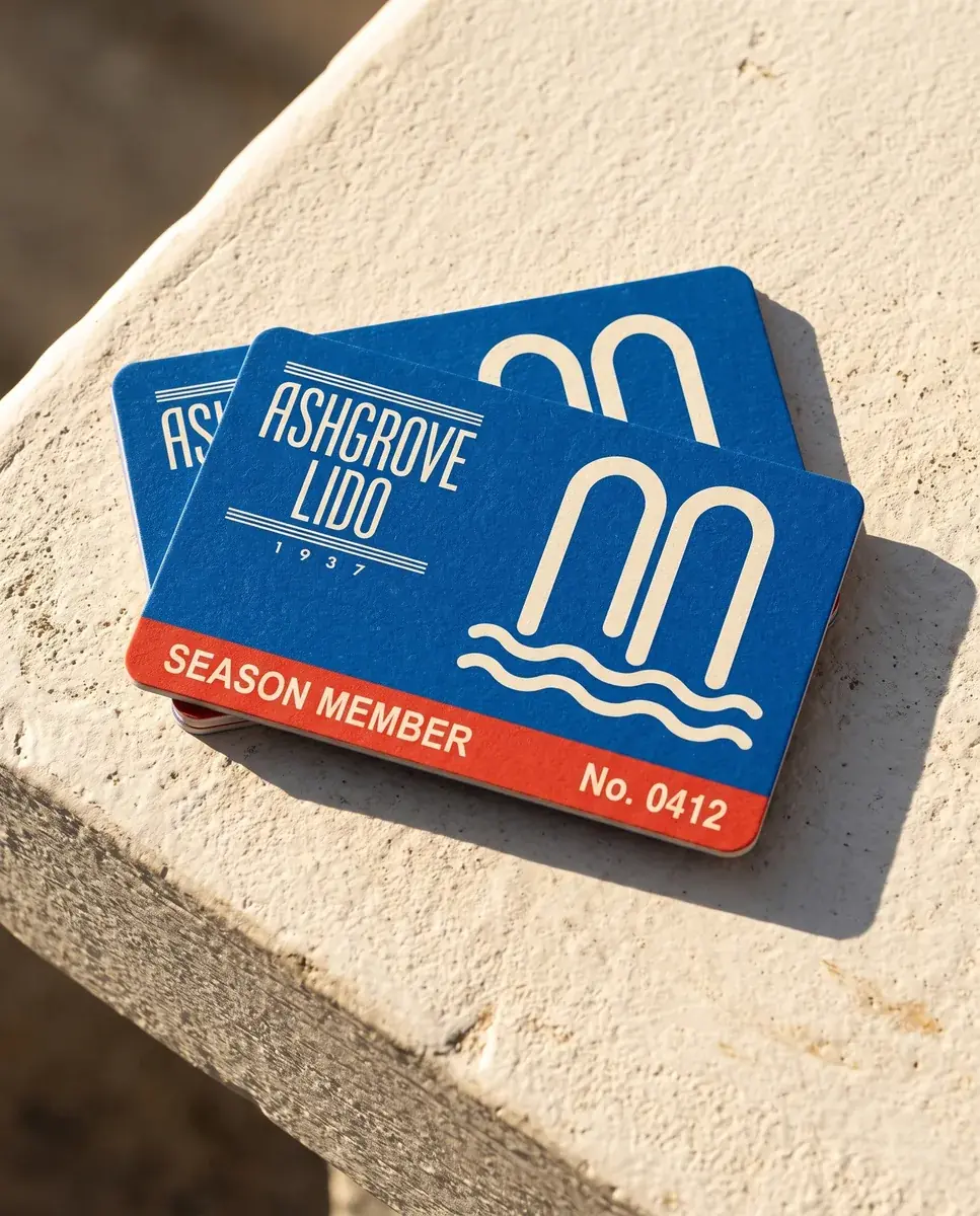 Two blue Ashgrove Lido membership cards fanned on a white stone ledge in hard sun, the wordmark and the arch mark in cream, a red band reading SEASON MEMBER and No. 0412
