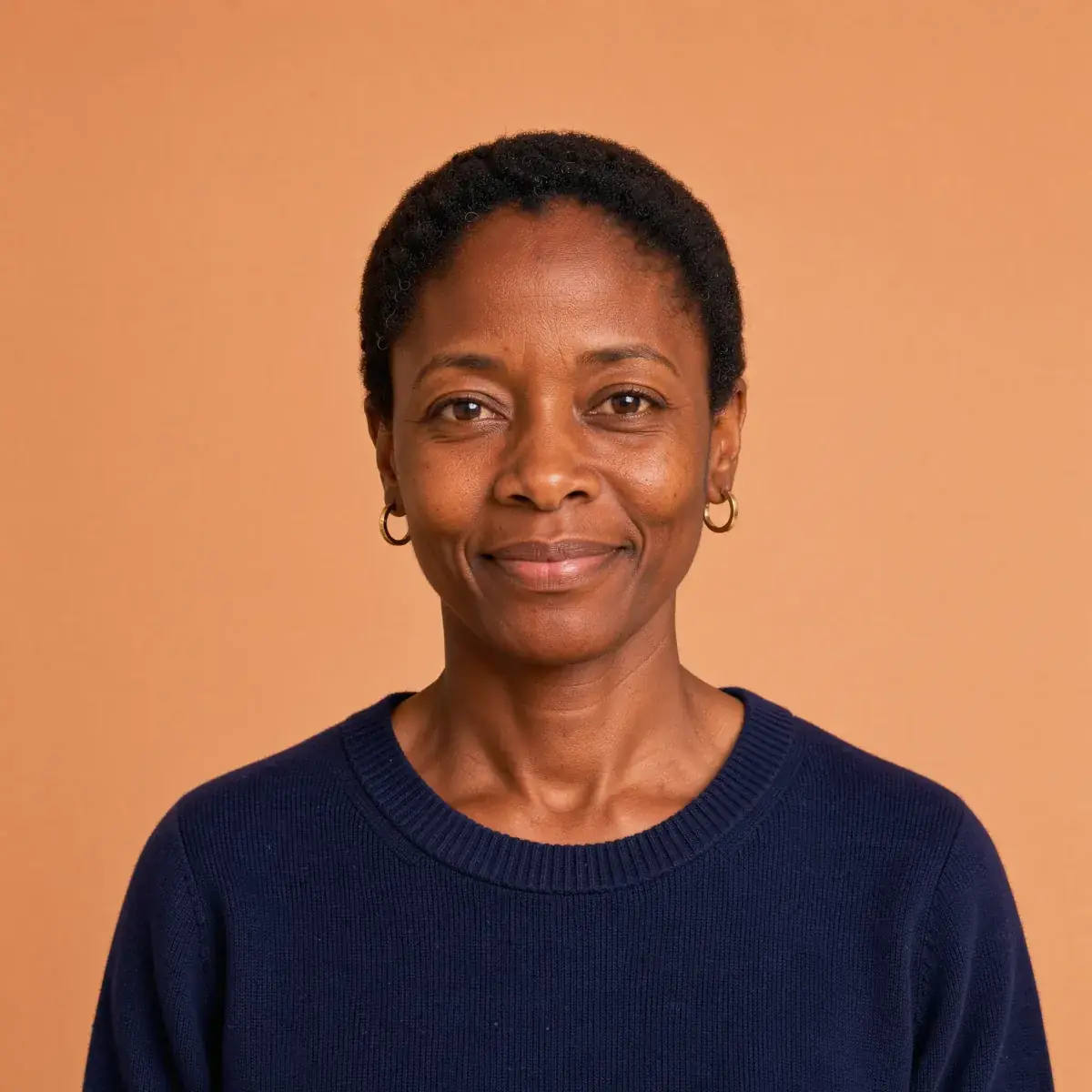 A woman with close-cropped hair, gold hoops and a navy sweater against an even peach backdrop, natural skin, made on Flux 3 Image