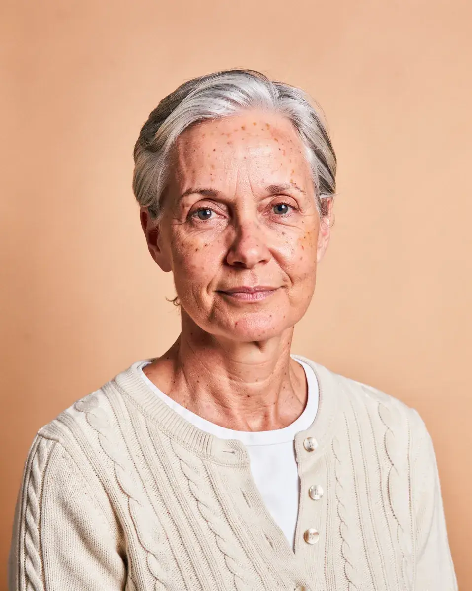 The next portrait in the set: a woman in her early sixties with short swept-back silver hair and a few freckles, in a cream cable-knit cardigan over a white collarless shirt, on the same peach backdrop and light