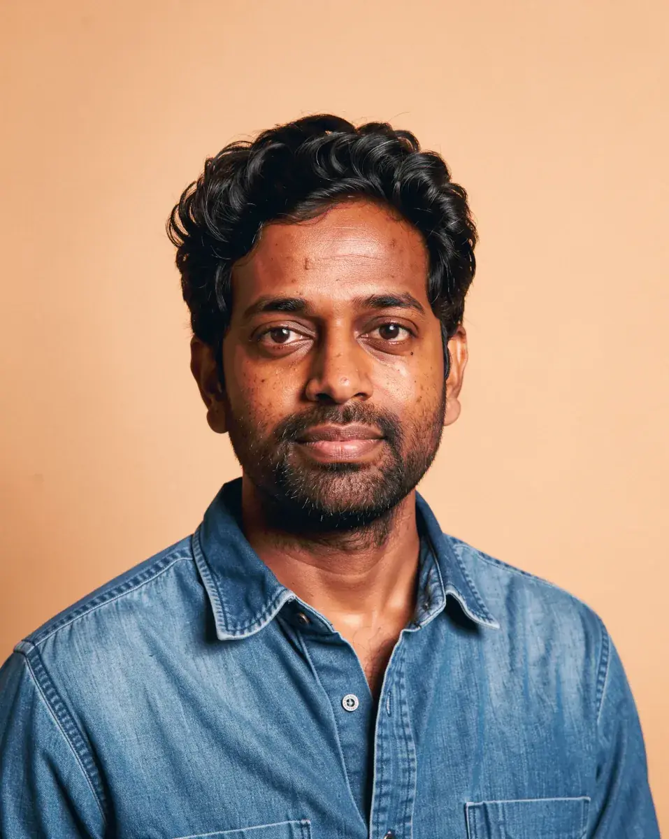 The next portrait in the set: a man in his mid thirties with dark wavy hair and a short trimmed beard, in a washed denim shirt, on the same peach backdrop and light