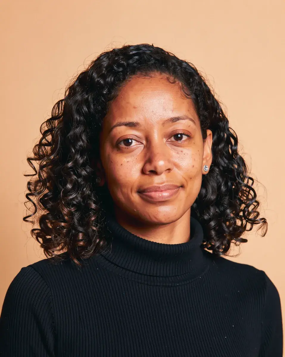The next portrait in the set: a woman with dark shoulder-length curls and small silver studs, in a black ribbed turtleneck, on the same peach backdrop and light