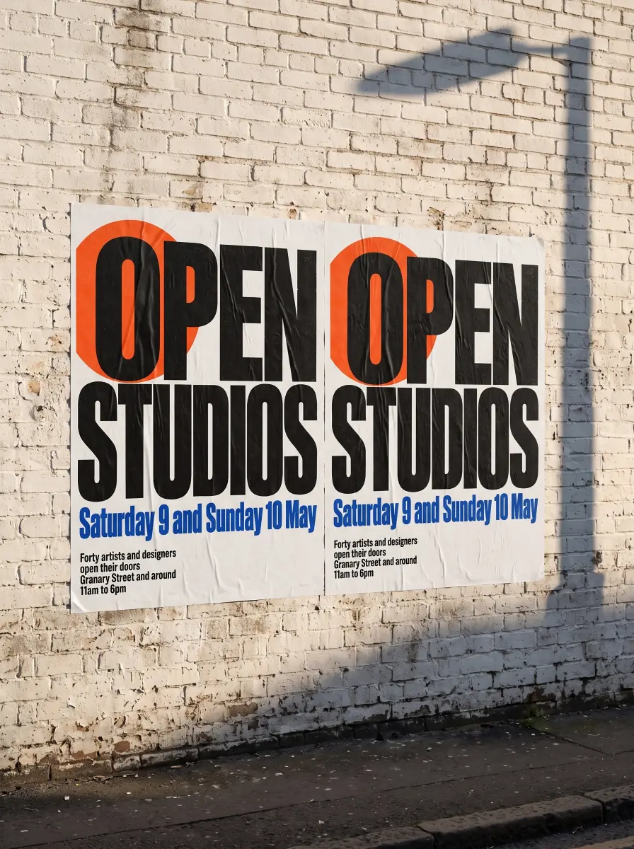 Two copies of the open studios poster pasted side by side on a whitewashed brick wall in late afternoon sun, the shadow of a street lamp across the wall