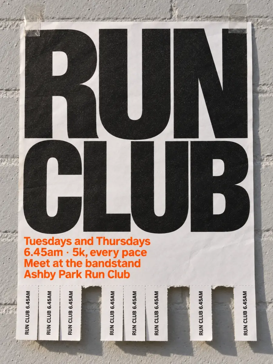 A white flyer taped to a grey painted brick wall: RUN CLUB in huge black capitals, the days, time, meeting point and Ashby Park Run Club in orange, and a row of RUN CLUB 6.45AM tear-off tabs with three torn away