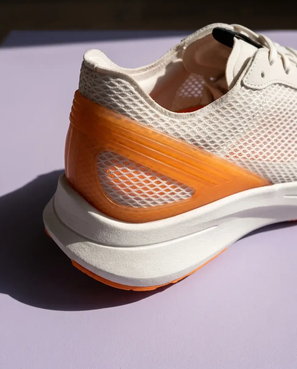 A close-up of the ribbed orange heel clip over the white mesh and the midsole edge, on a pale lilac plane in raking sun