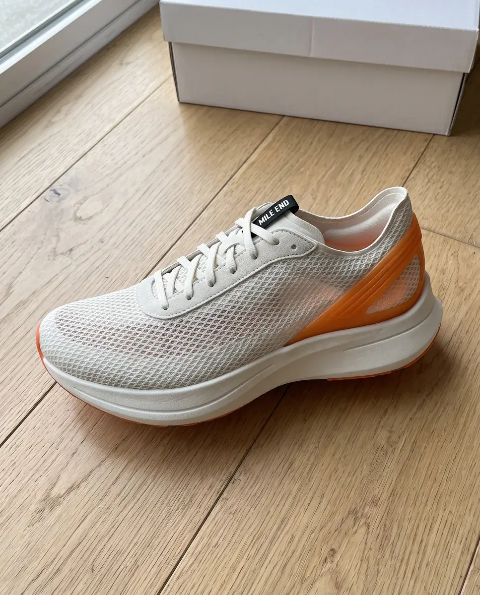 A generated phone photo of a white mesh running shoe with an orange heel clip on a light oak floor, a white shoe box lid at the top edge