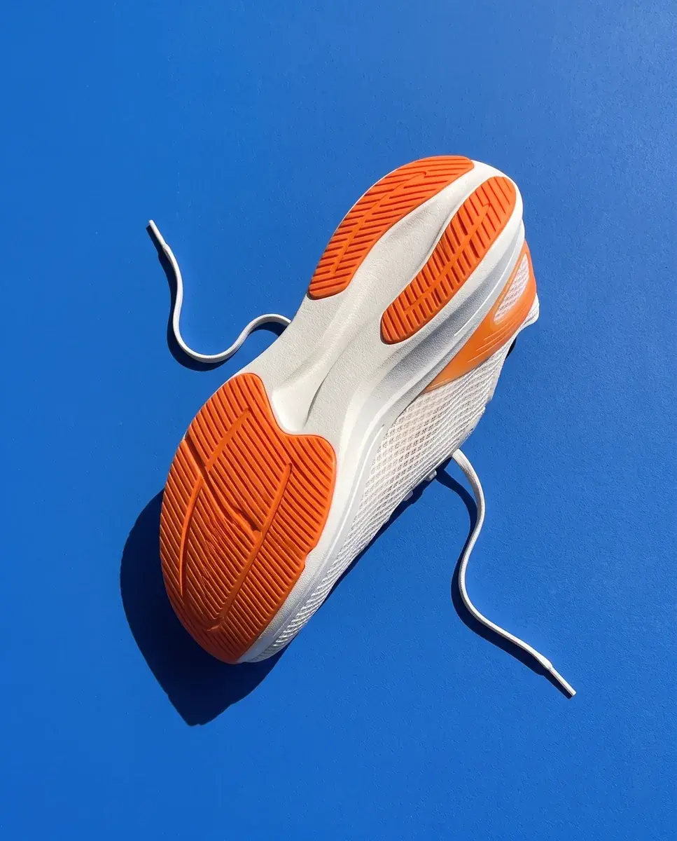 The shoe turned over on a cobalt plane, seen from above: two grooved orange outsole pads and white foam, the laces curving loose across the blue