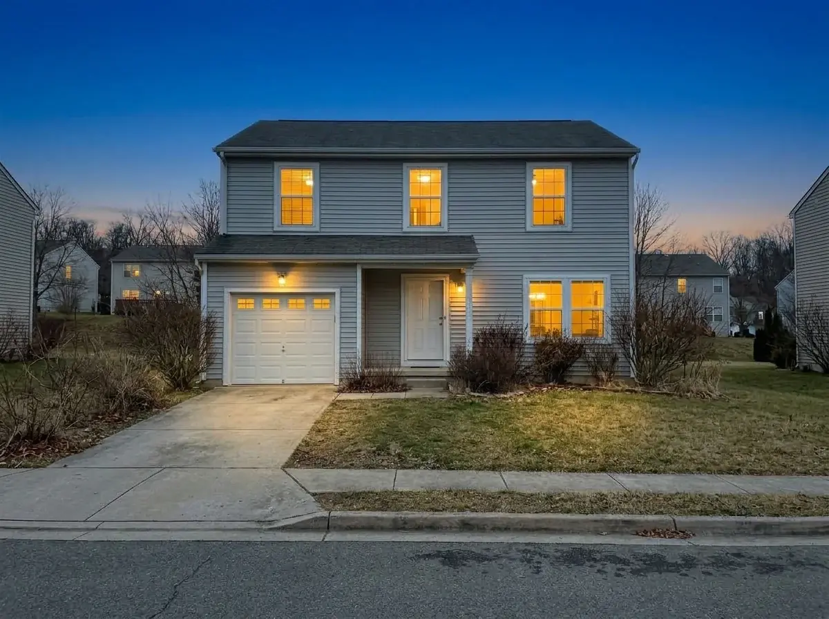 The same house edited by AI to twilight: a deep blue dusk sky with an orange glow, warm light in every window and the porch and garage lights on