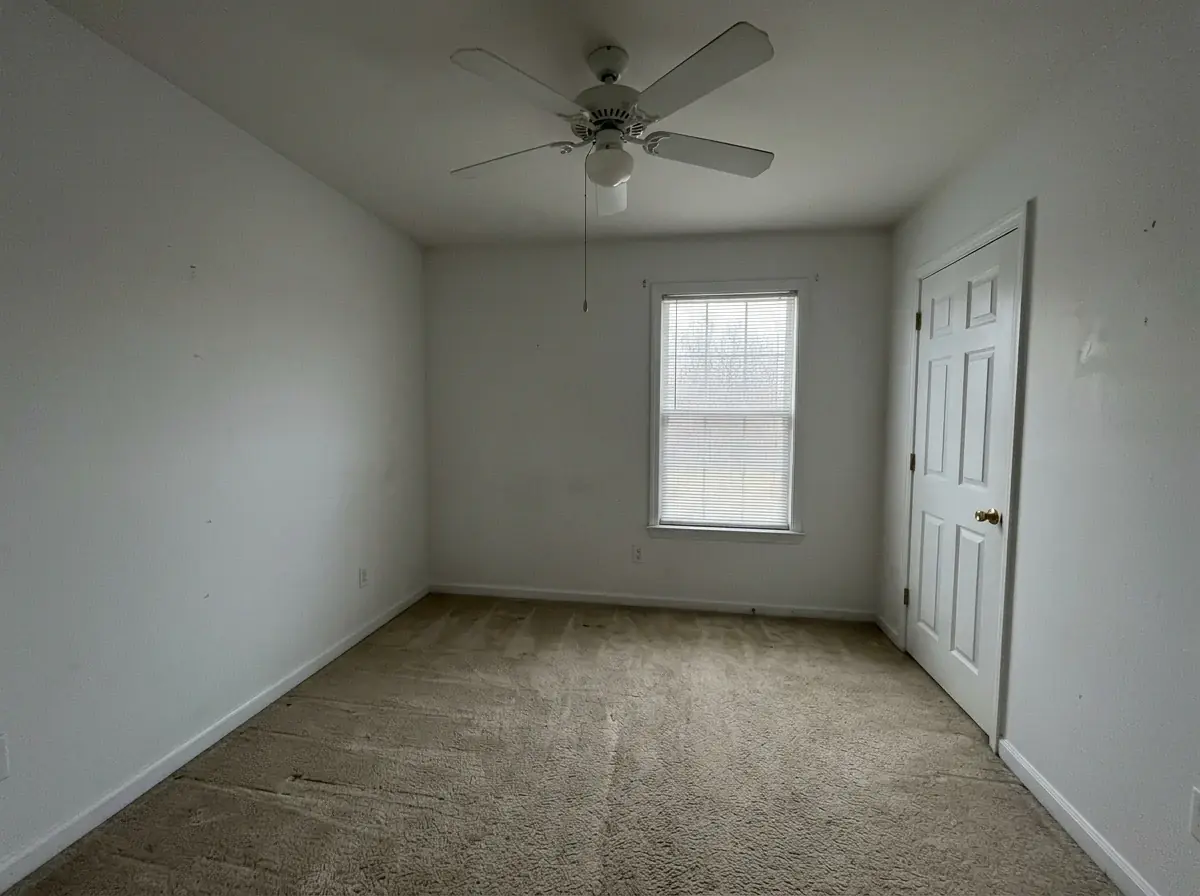 A generated phone photo of an empty spare bedroom: white walls, beige carpet, a ceiling fan, a closet door and one window with blinds, in flat grey light