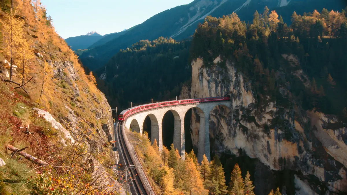 A red train on a curved stone viaduct heading into a cliff tunnel, seen from a steep slope of golden larches in warm low sun, made on Flux 3 Image