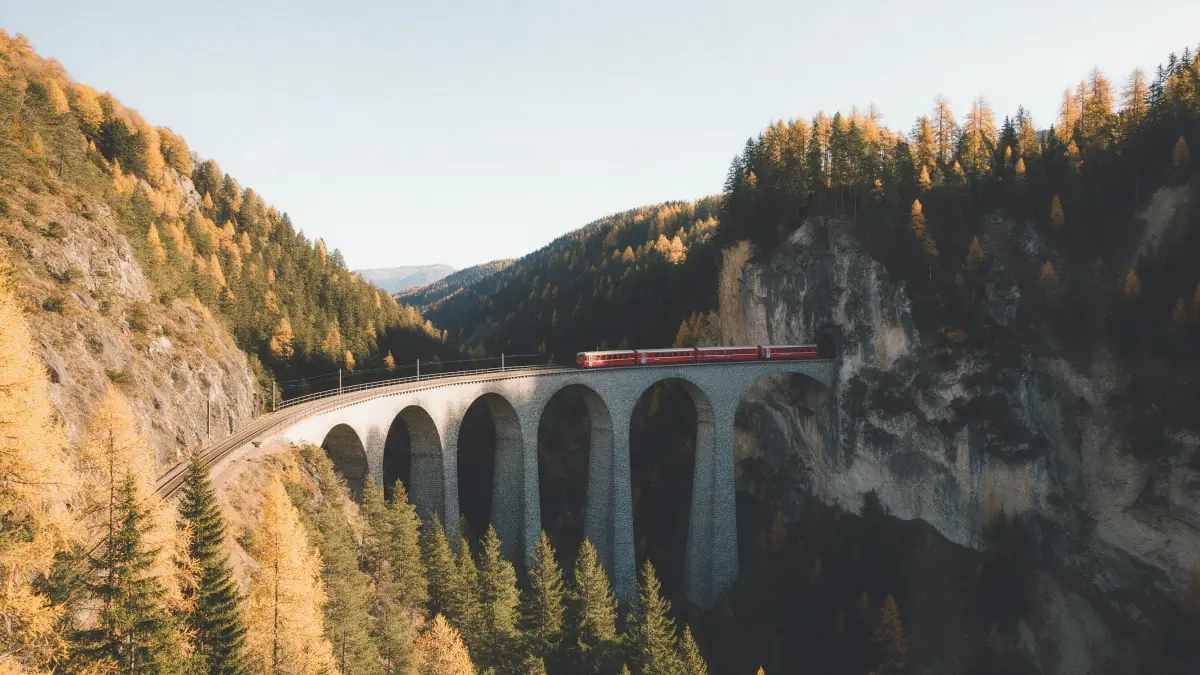 A red train on a tall arched viaduct between larch slopes and a cliff, open pale sky above, in a faded film grade, made on Seedream 5 Pro