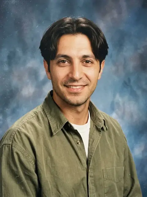 A 90s yearbook portrait of a man with a curtain haircut in an olive shirt over a white tee against a mottled blue backdrop
